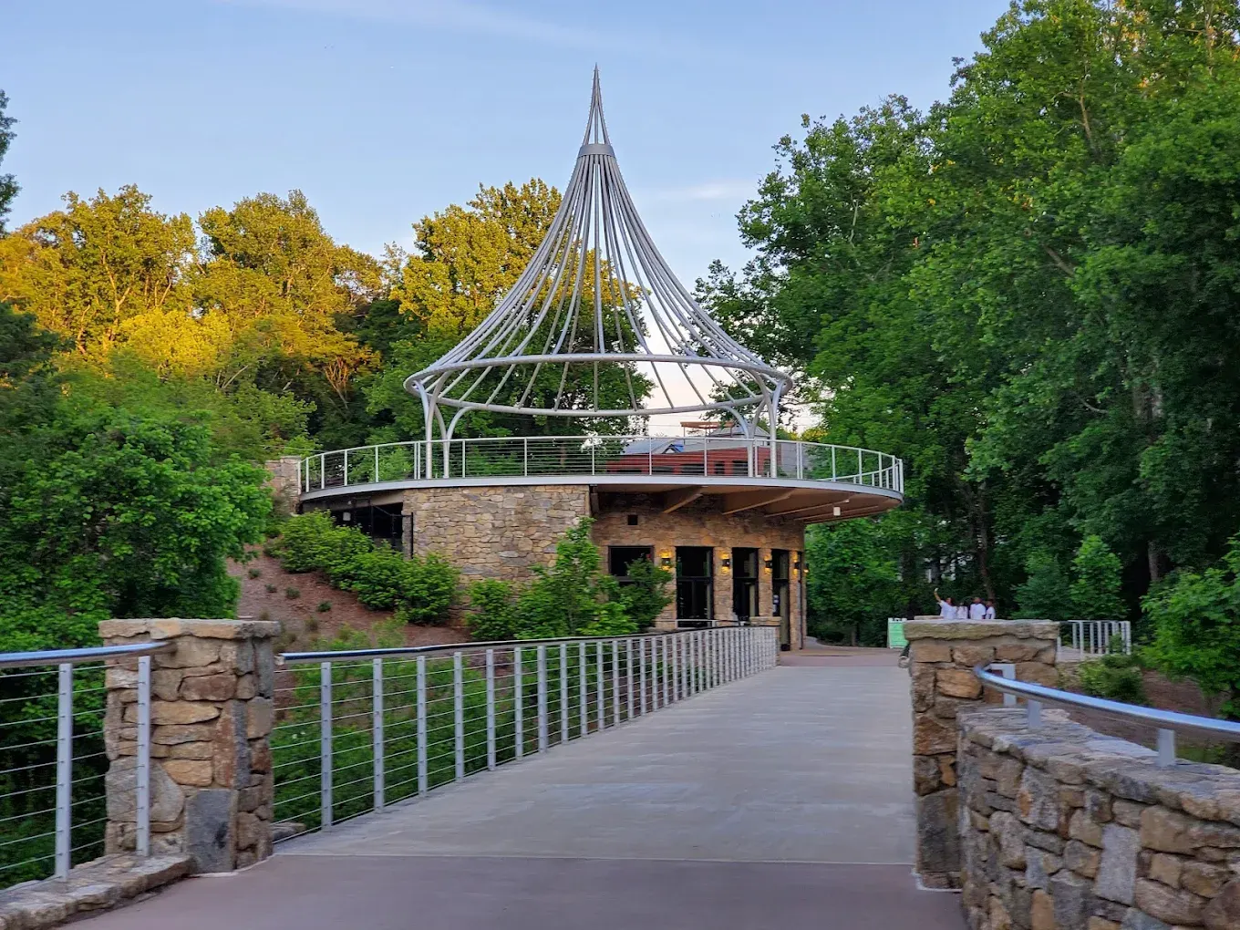 A paved pedestrian bridge leading to a round stone building topped by a tall, tent-like silver spire, framed by green trees, at Cancer Survivors Park Center for Hope & Healing.