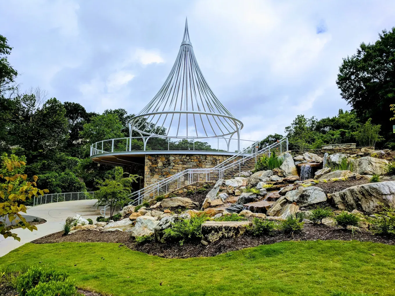 The pavilion at Cancer Survivors Park Center for Hope & Healing: a round stone building topped by a tall, tent-like silver spire, beside a terraced rock-and-water garden and green lawn.